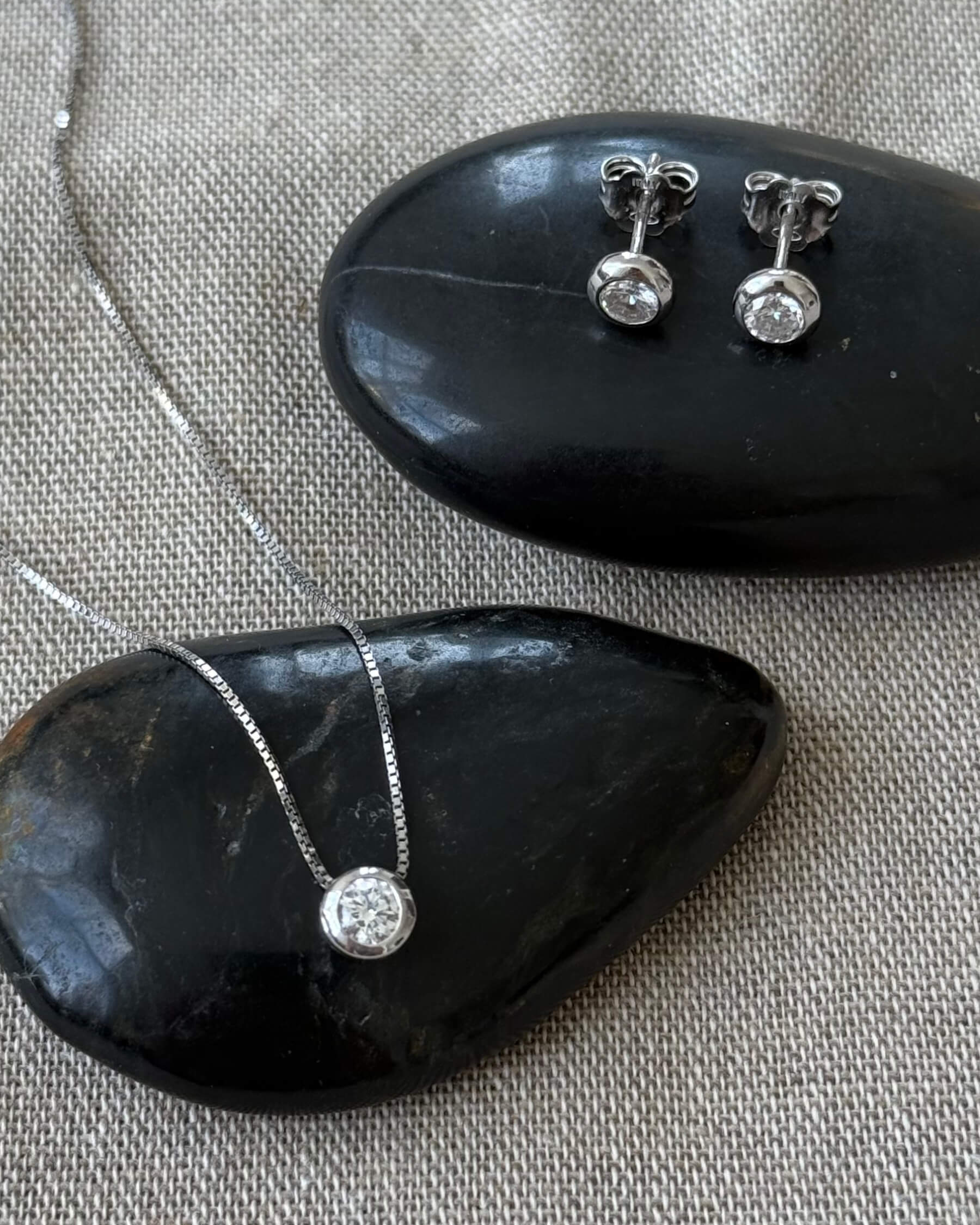 Halsband med tunn kedja och runt hängsmycke med en gliittrande CZ (genomskinlig glaskristall) och örhängen med CZ (genomskinlig glaskristall) i rhodiumpläterat 925 sterling silver. Halsbandet och örhängena ligger på en varsin svart sten.