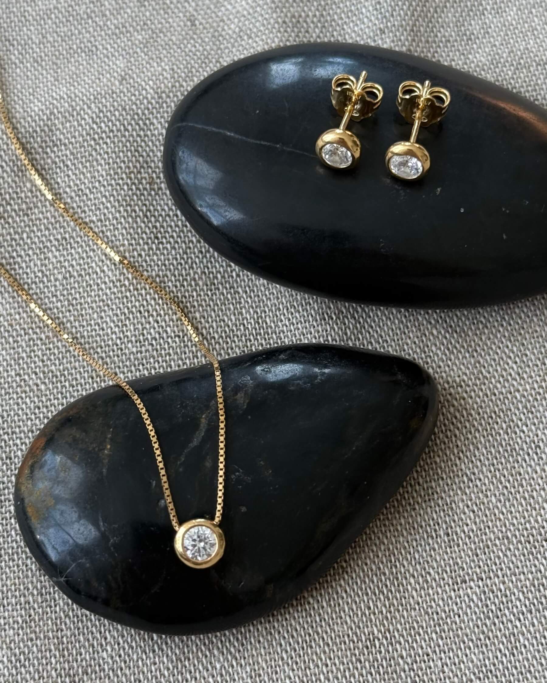 Halsband med tunn kedja och runt hängsmycke med en gliittrande CZ (genomskinlig glaskristall) och örhängen med CZ (genomskinlig glaskristall) i guldpläterat 925 sterling silver. Halsbandet och örhängena ligger på en varsin svart sten.