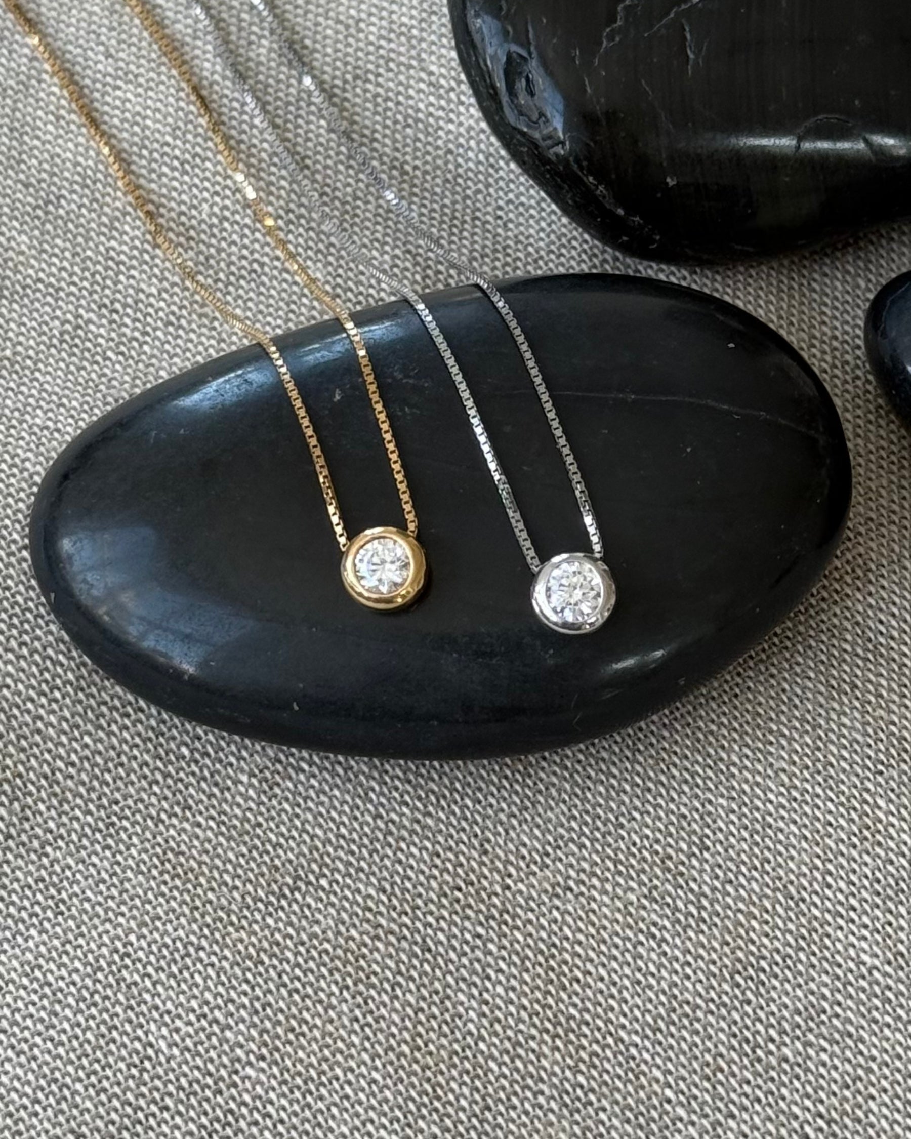Två halsband med tunn kedja och runt hängsmycke med en gliittrande CZ (genomskinlig glaskristall) som ligger på en svart sten. Halsband till vänster i guldpläterat 925 sterling silver och halsband till höger i rhodiumpläterat 925 sterling silver.