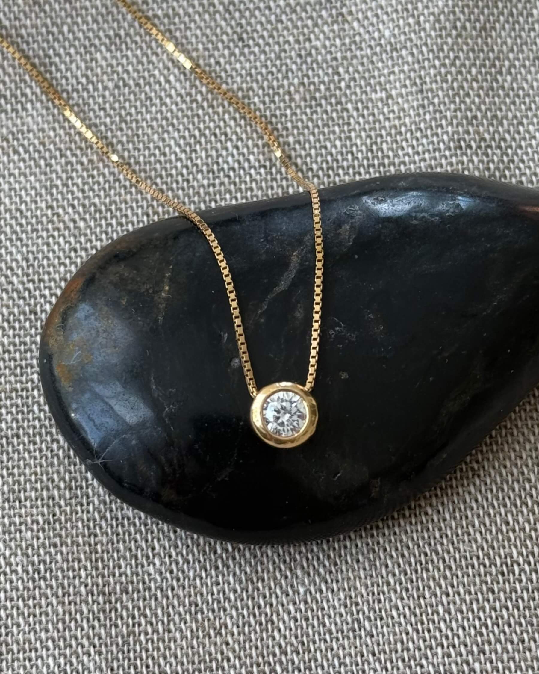 Halsband med tunn kedja och runt hängsmycke med en gliittrande CZ (genomskinlig glaskristall) i guldpläterat 925 sterling silver. Halsbandet ligger på en svart sten.