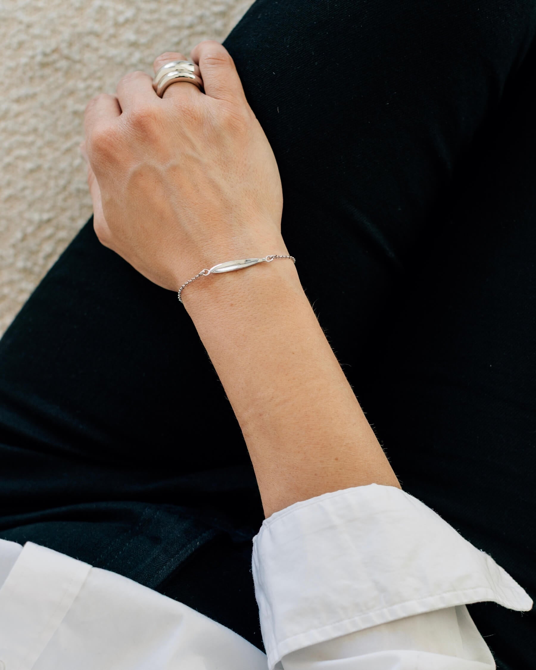 Arm liggandes i knä som bär ett elegant armband med tunn kedja och en blank olivbladsformad platta, tillverkat i rhodiumpläterat 925 sterling silver.