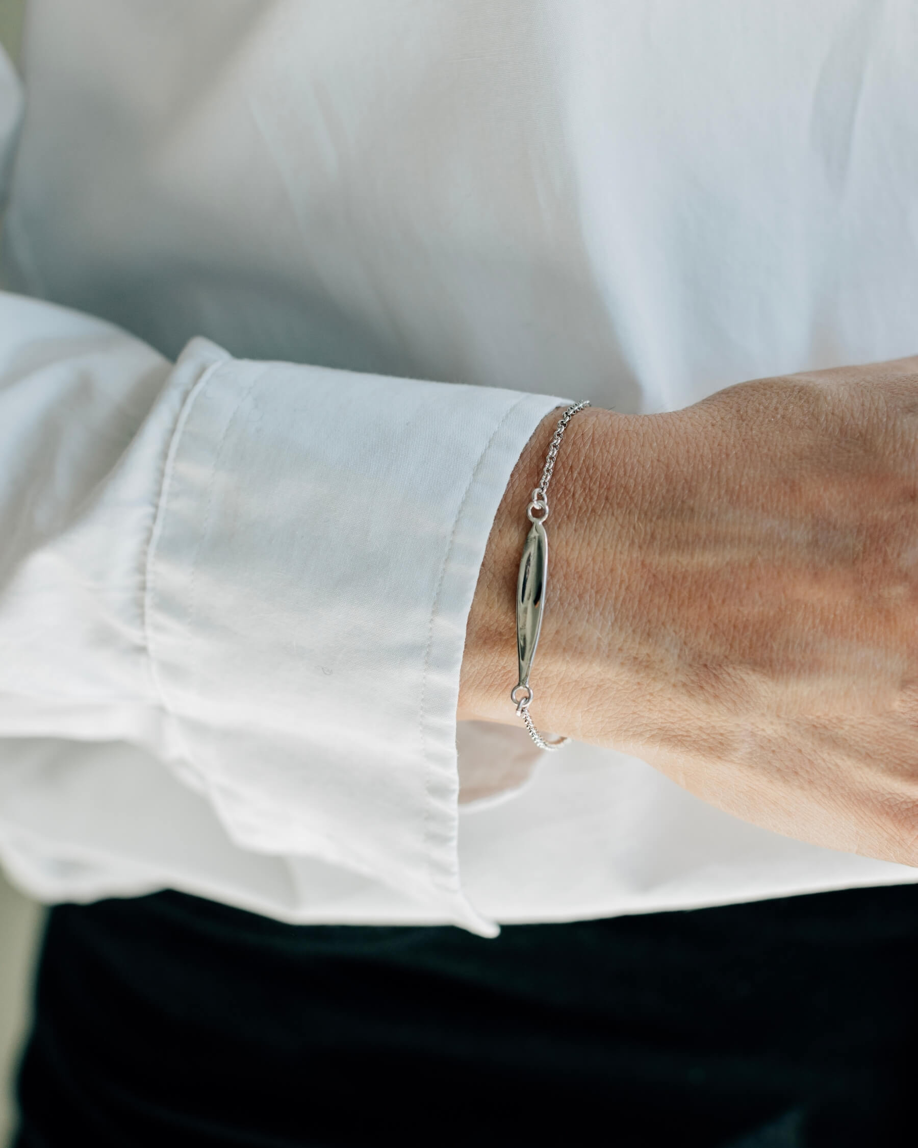 Närbild på en handled som bär ett elegant armband med tunn kedja och en blank olivbladsformad platta, tillverkat i rhodiumpläterat 925 sterling silver.
